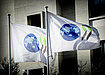 Two white flags, each with a blue globe and two black and green chevrons printed on them, blow in the wind in front of the OECD Paris headquarters building.