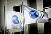 Two white flags, each with a blue globe and two black and green chevrons printed on them, blow in the wind in front of the OECD Paris headquarters building.