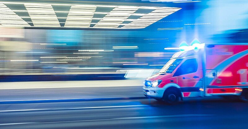 A red ambulance with sirens blaring rushing down a street in a blur. Photo by Camilo Jimenez via Unsplash.