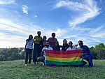 Hertie School Pride Network members hold up a rainbow flag on a green field under a bright blue sky.