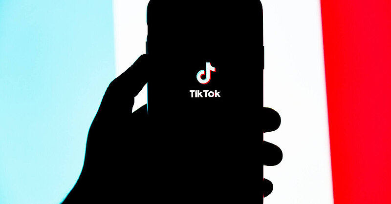 A silhouette of a hand holding up an iPhone with the TikTok logo illuminated on screen. Photo by Solen Feyissa via Unsplash.