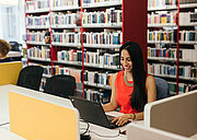 Student in a bright red-orange dress with long black hair, seated in a library in front of a black laptop. Rows of bookshelves are in the background.