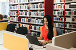 Smiling person in red dress looking at laptop, surrounded by shelves of books