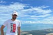 Danial stands atop the Bokor Mountains overlooking a scenic landscape with sweeping blue skies in Kampot, Cambodia. He wears a white baseball cap with the red letters "giz" on it.