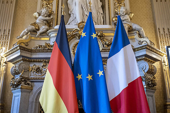The German and French flags flank the blue European Union flag on the left and right, respectively, on a blue stage in a gilded and lavishly decorated hall.