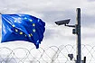 Photo of EU flag behind a barbed wire fence and surveillance camera at EU border