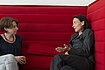 Two people sitting with crossed legs and conversing on a red couch.
