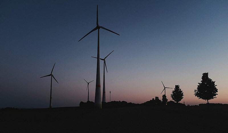 Silhouettes of a wind farm and some trees against a dark blue-orange sky.