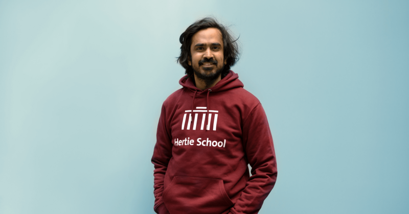 Sanyam stands against a blue background, wearing a maroon hoodie with a white Hertie School logo on it.