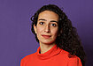 Caroline in a bright red-orange turtleneck in front of a purple background.