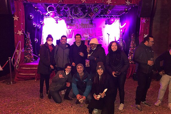 The Hertie School Pride Network gather at a queer Christmas Market in Berlin. They are bundled in warm clothing and purple lights shine behind them.