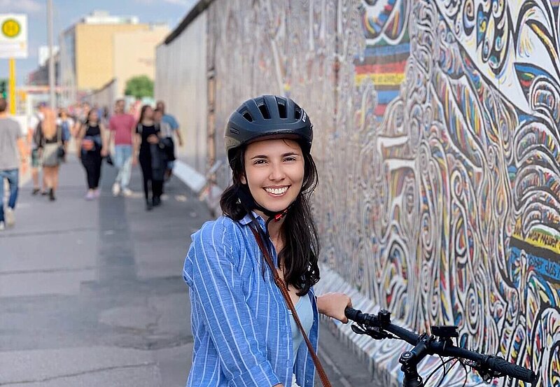Carol wearing a helmet and standing in front of the Berlin East Side Gallery with a bike.