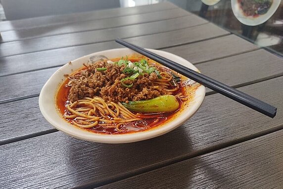 A bowl of spicy noodles in red oily sauce sits on a table with a pair of chopsticks placed to the side.